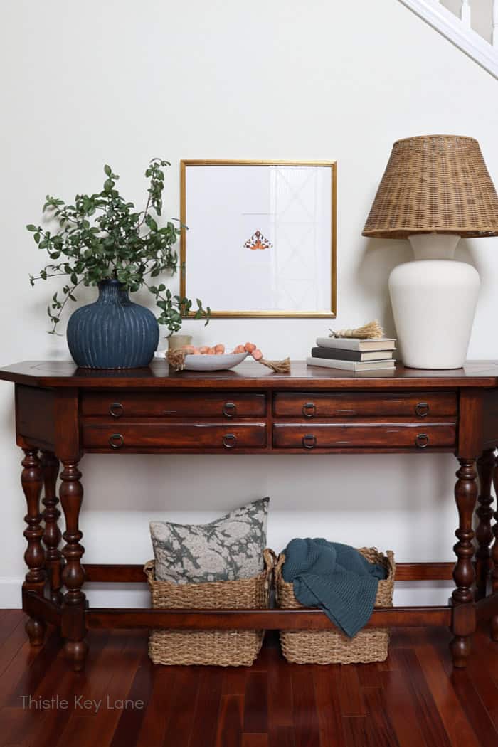 Entry table with lamp, frame, vase, greenery, baskets below.