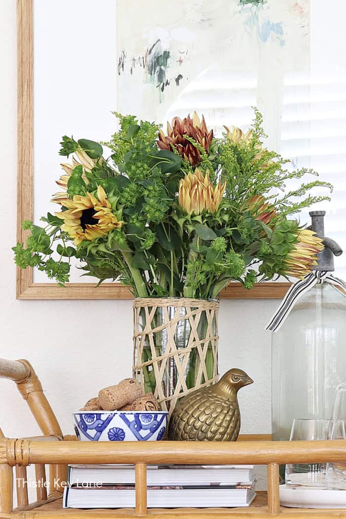 Vase of fresh sunflower and protea flowers with greenery on a wooden shelf in home decor arrangement. Summer To Fall Bar Cart With Sunflowers.