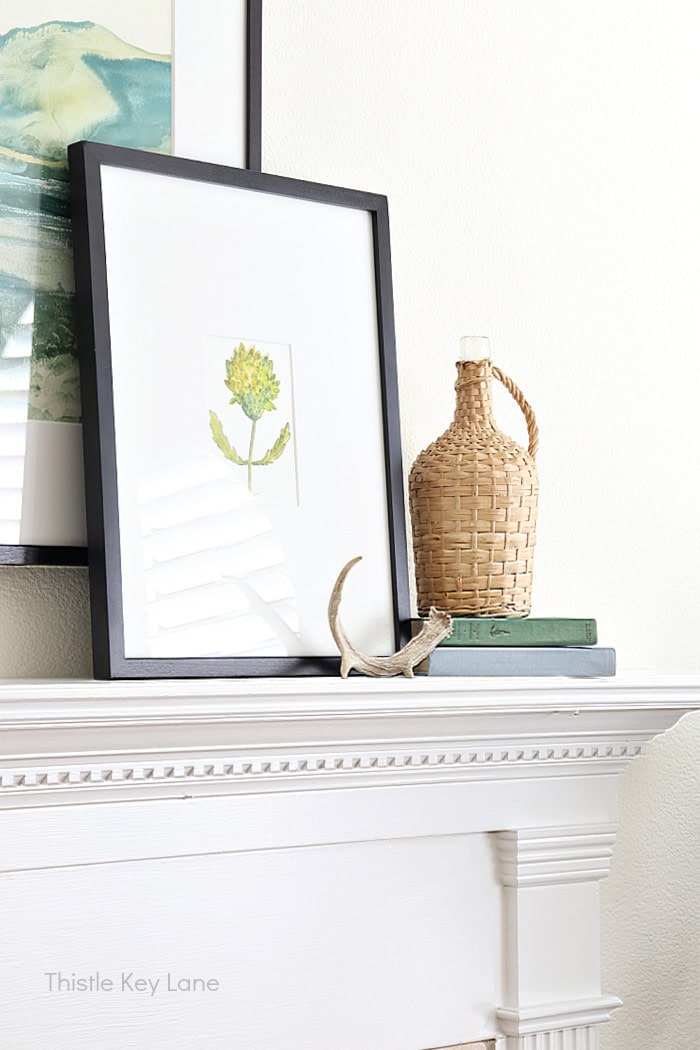 Art print with green and yellow floral watercolor artwork on black frame, wicker wine bottle, deer shed, and books on white mantel.