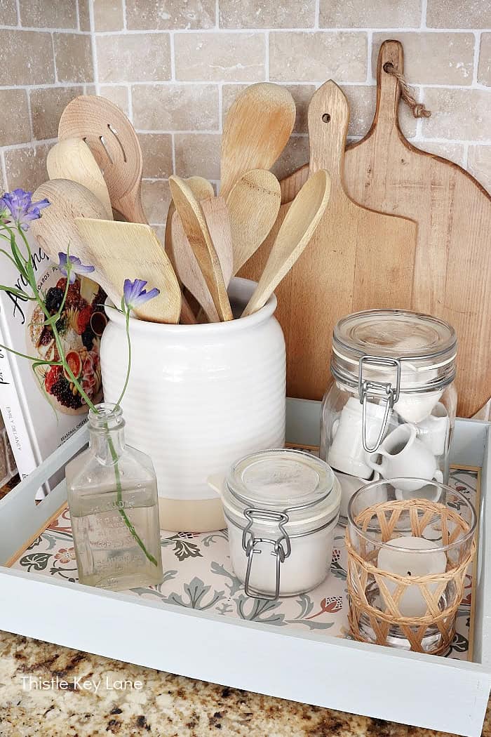 Wood tray on counter with jars, crock, candle and cutting boards. How To Decorate A Tray With Wallpaper.