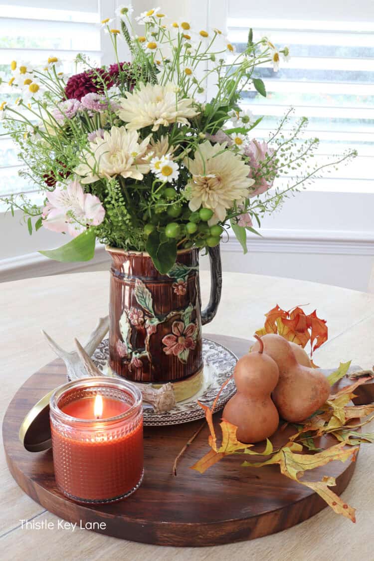 Fall arrangement with candle. <!-- wp:image {"id":35414,"sizeSlug":"large","linkDestination":"none","pinterestText":"Fall Floral Centerpiece With Vintage Pitcher. See how to create a little vignette for your kitchen table or island featuring a vintage pitcher, pretty flowers and seasonal accents. Fall vignette with vintage majolica pitcher.","pinterestTitle":"Fall Floral Centerpiece With Vintage Pitcher - Thistle Key Lane"} -->
<figure class="wp-block-image size-large"><img src="https://thistlekeylane.com/wp-content/uploads/2026/10/majolica-pitcher-flower-arrangement-3-750x1125.jpg" alt="Bottleneck gourds with fall leaves. Fall Floral Centerpiece With Vintage Pitcher. Fall Floral Centerpiece With Vintage Pitcher.
